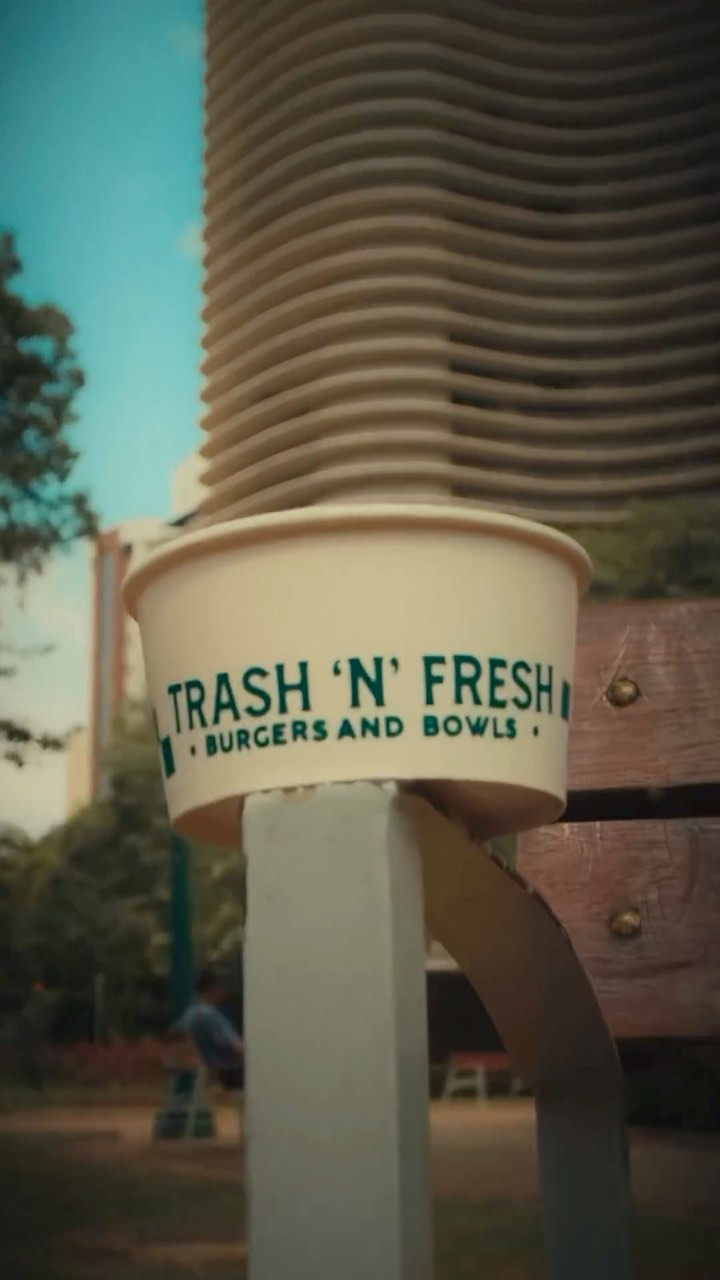 Copo de papel creme com o logo Trash 'n' Fresh em verde, apoiado num banco de praça com um prédio de linhas curvas ao fundo