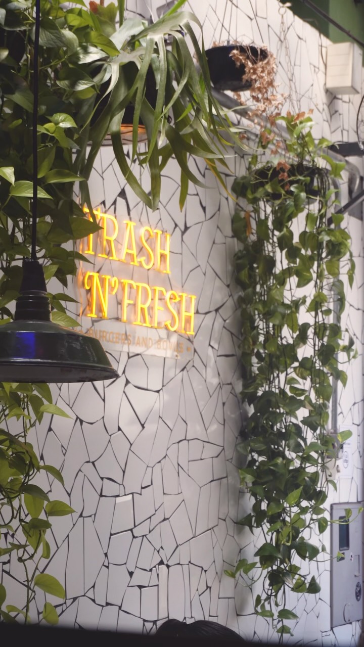 Letreiro neon amarelo Trash 'n' Fresh na parede de mosaico de cacos brancos, entre plantas pendentes e uma luminária de metal