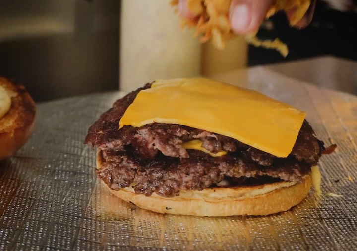 Smash duplo na chapa com cheddar derretendo, recebendo crispy de cebola
