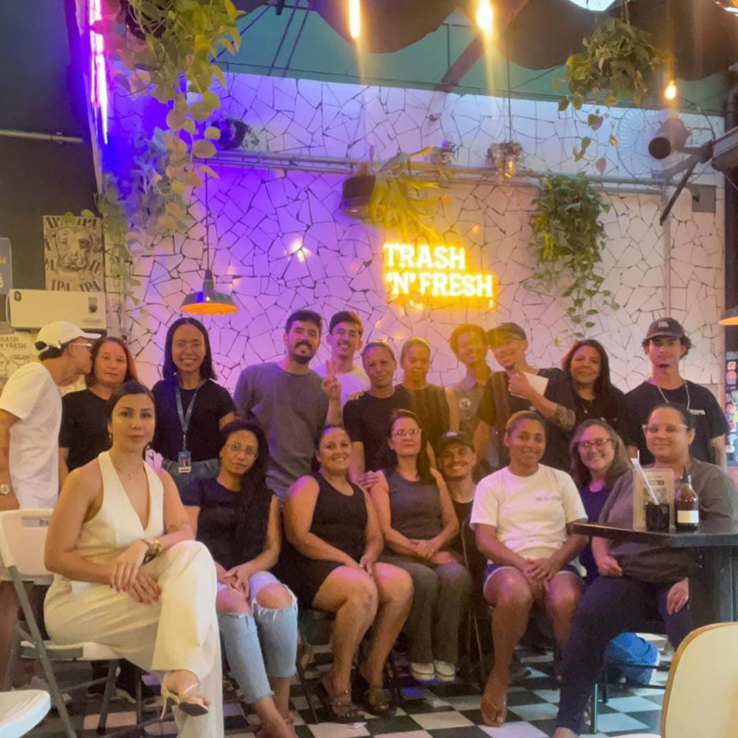 A equipe da Trash 'n' Fresh reunida no salão, embaixo do neon e das plantas, sobre piso xadrez