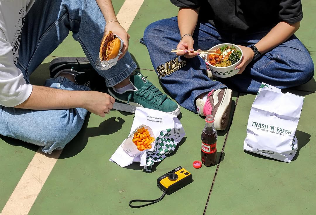 Duas pessoas sentadas numa quadra verde: uma com burger embrulhado no papel alumínio, outra com poke bowl e hashis, sacola da Trash 'n' Fresh e batatinha ao lado