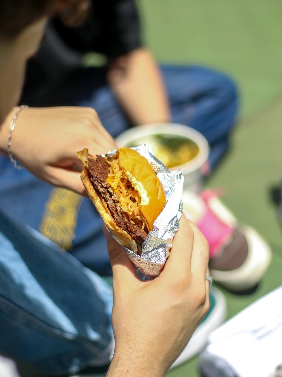 Mão segurando um smash burger embrulhado no papel alumínio, com um bowl ao fundo