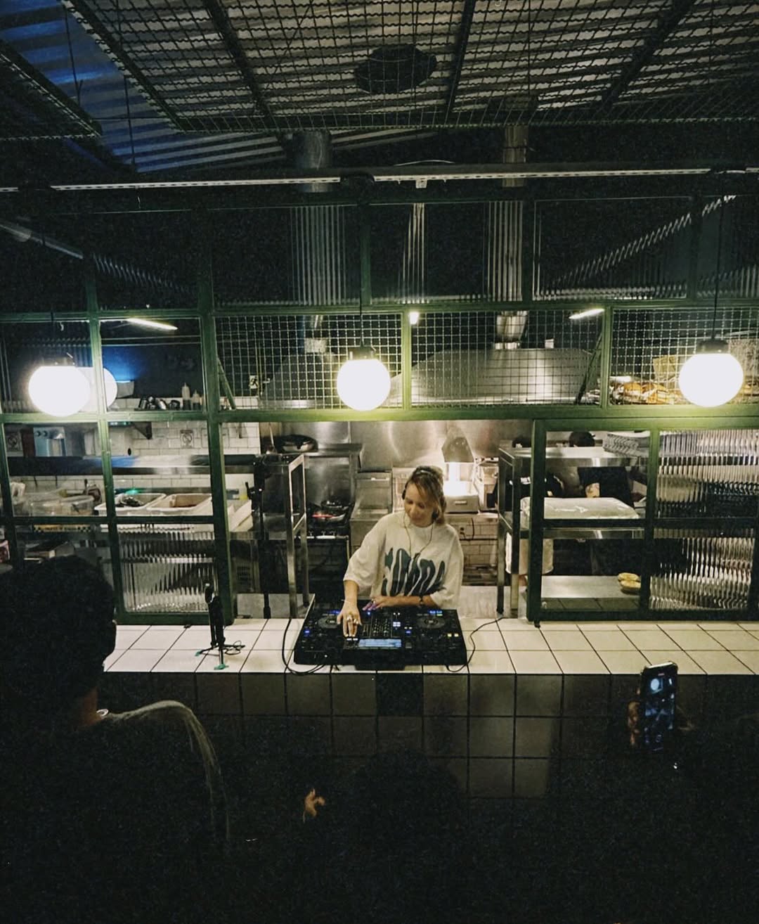 DJ tocando house no balcão de azulejos brancos, com a cozinha de vidro e luminárias redondas ao fundo