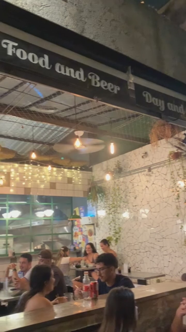 Salão cheio à noite, com luzinhas, mosaico branco e plantas; placa Food and Beer no alto
